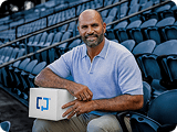 Albert Pujols sentado en las gradas de un estadio con una caja Cologuard en su regazo.